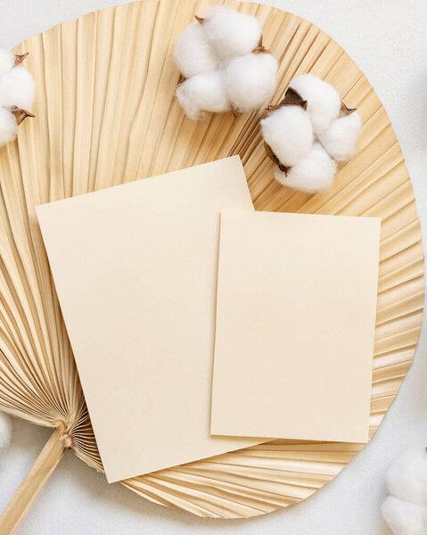 Blank cards on dried palm leaf with cotton flowers top view, mockup. Romantic scene with vertical paper cards. Bohemian pastel greeting or wedding Invitation