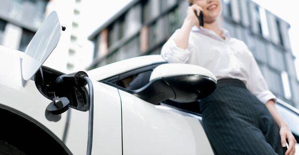 Focus charging-electric car with EV charger at charging station with blur businesswoman talking on phone with residential building in background as progressive lifestyle concept.