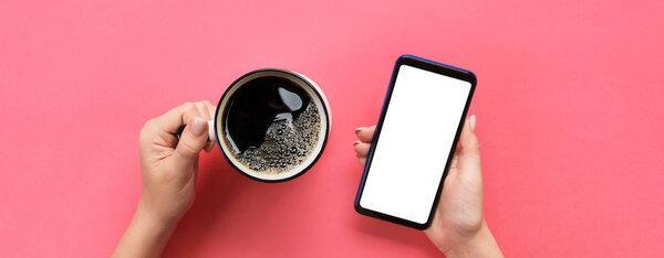 Female hands holding black mobile phone with blank white screen and mug of coffee. Mockup image with copy space for you design. Top view banner on pink background, flat lay.
