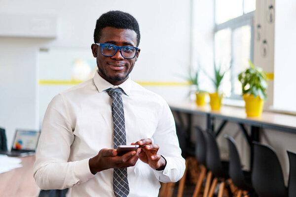 Confident, smiling male engages with mobile business app, connected and tech-savvy. Pro African businessman in shirt and tie uses smartphone in modern office setting.