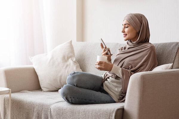 Modern arabic girl sitting on couch with smartphone and drinking coffee