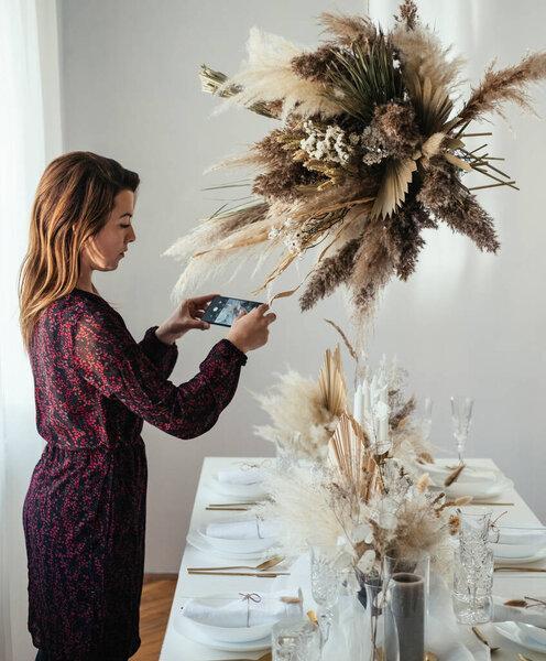 Young Woman Taking a Picture of Decorated Christmas Dinner Table at Home. Elegant female in dress photographing table set up in dinning room with her mobile phone before weeding lunch party.