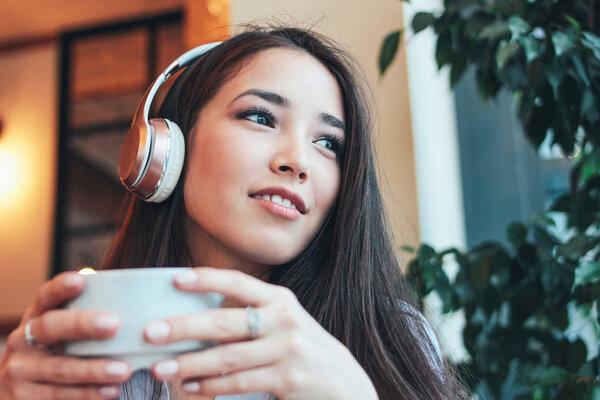 Beautiful charming brunette smiling Asian girl enjoys music in headphones with cup of coffee or tea in cafe