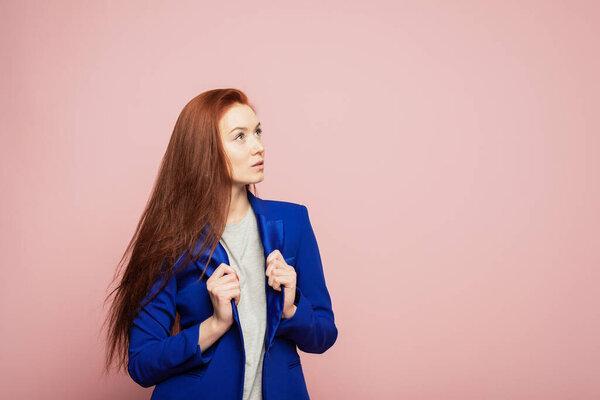 A redhead girl is on the pink background. A thoughtful redhead girl is on the pink background. A girl is in blue jacket on the pink background. A girl with beautifull red hair on the pink background. A girl is looking into the distance.
