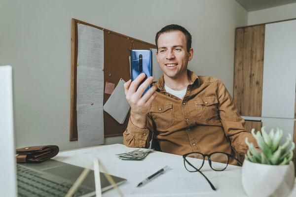 Young man using smartphone sitting at the desk in the room indoor. Communication online, video chat. Freelancer using technology at home. Selective focus.