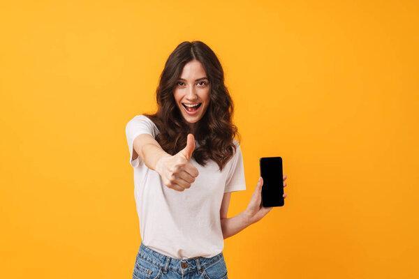 Happy positive smiling young woman posing isolated over yellow wall background using mobile phone showing display and thumbs up.