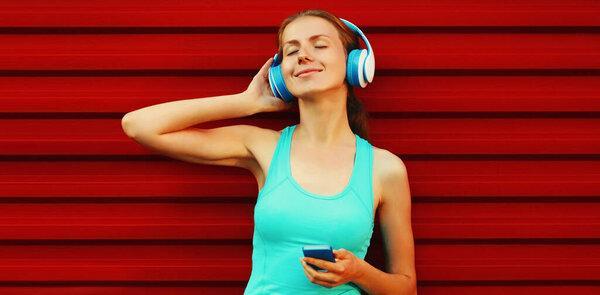 Portrait of fitness smiling young woman in headphones listening to music with smartphone on red background