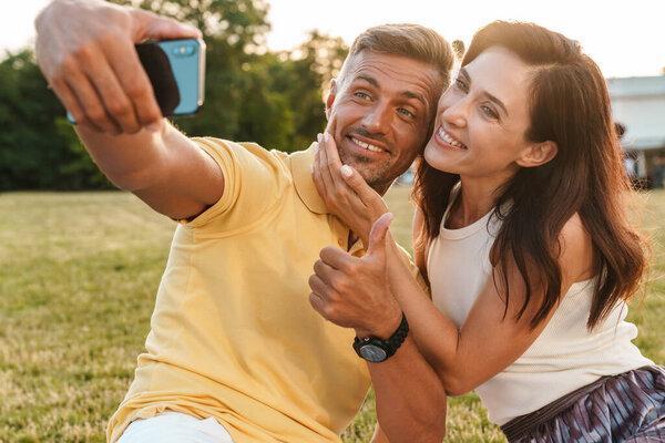 Portrait of pleased adult couple man and woman showing thumb up and taking selfie photo on cellphone while sitting on grass in park
