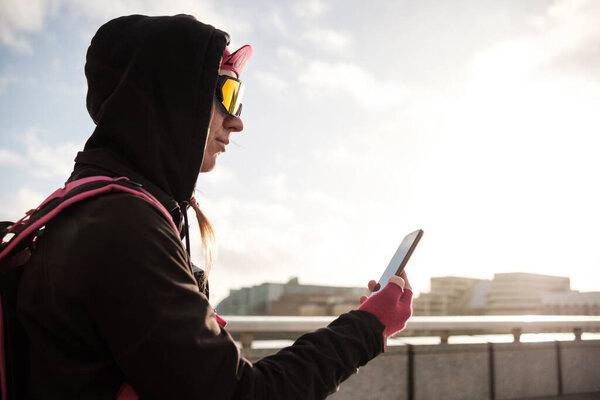 Side view of bike courier woman using her phone on London Bridge. She is wearing cycling outfit and sunglasses. She is looking at the screen.