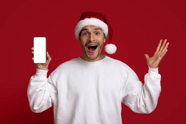 Amazing Offer. Excited Guy In Santa Hat Holding Smartphone With Blank Screen