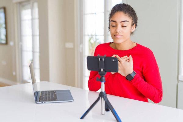 Young african american woman doing video call using smartphone camera smiling with hands on chest with closed eyes and grateful gesture on face. Health concept.