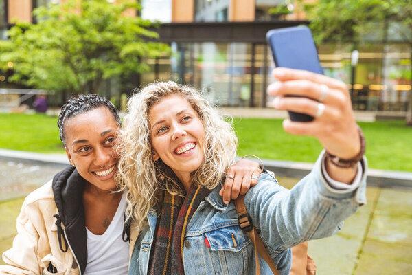 Happy mixed race girlfriends taking a selfie and kissing - Lesbian couple, millennials women, having fun together in London - LGBTQ concept with multiracial beautiful couple