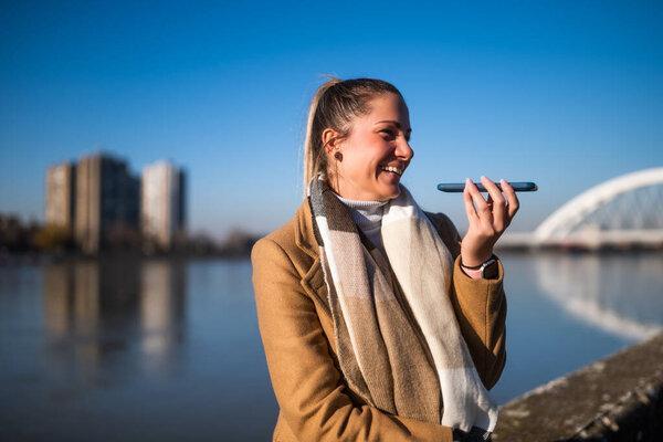 Beautiful woman in warm clothing talking on a mobile phone and enjoys resting by the river on a sunny winter day.
