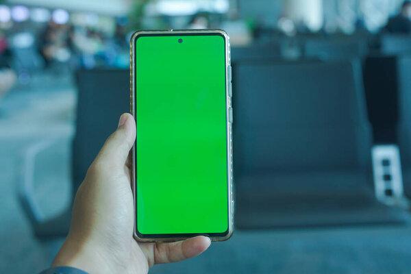 Person holding a smartphone with a green screen at an airport departure lounge