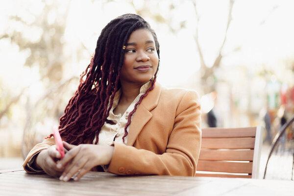 Young african american woman sitting in street cafe and holding smartphone in her hands