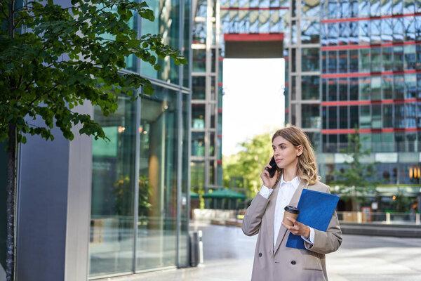 Corporate woman in business suit, walking in city center with work papers and mobile phone, calling someone.