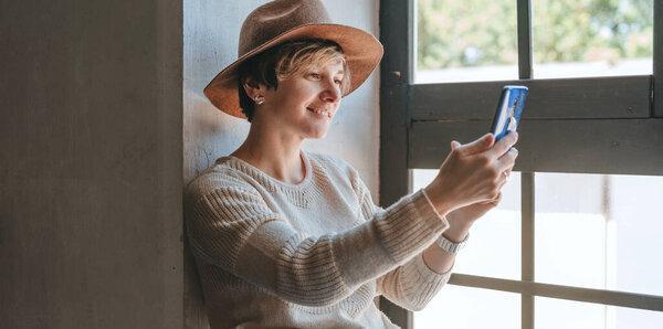 Young woman using smartphone having video call sitting near the window indoor. Online meeting or conference.