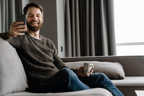 Bearded man using mobile phone and drinking coffee while sitting on sofa at home