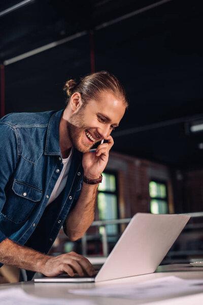 cheerful businesswoman using laptop while talking on smartphone