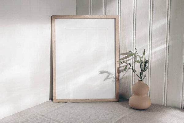 Portrait empty wooden frame mockup in sunlight. Olive branch in modern organic shaped vase. Beige linen table cloth. White wainscot wall paneling background. Scandinavian interior, home design. Art