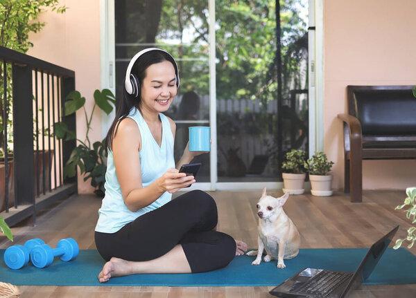 Portrait of Asian woman wearing headphones, holding coffee cup, using smartphone, sitting  on yoga mat in balcony  with computer laptop dumbbell and Chihuahua dog. Yoga exercise training online