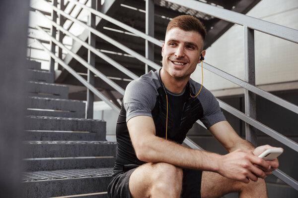Image of laughing caucasian sportsman using cellphone and earphones while sitting on urban stair outdoors