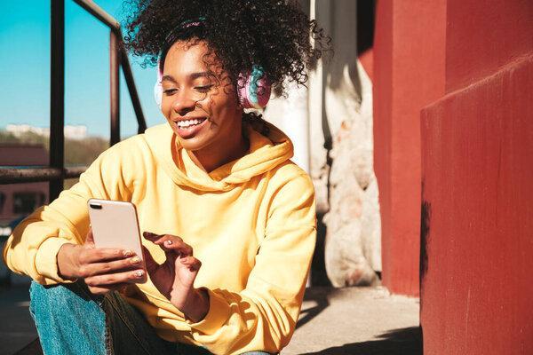 Beautiful black woman with afro curls hairstyle.Smiling model in yellow hoodie.Sexy carefree female enjoying listening music in wireless headphones.Posing on street background at sunset.Holds phone