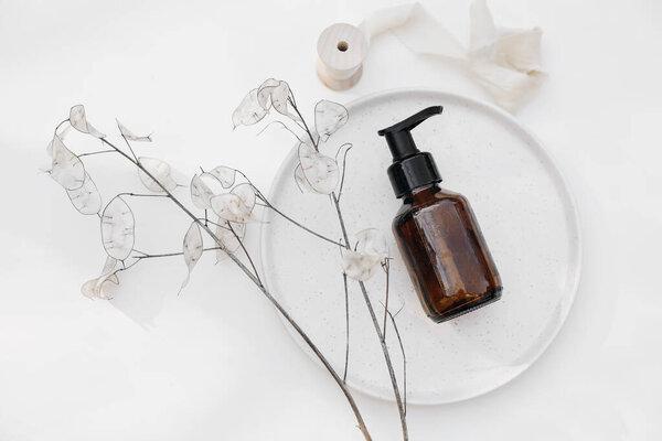 Amber glass cosmetic bottle. Shampoo pump dispenser on ceramic plate. Dry lunaria flower isolated on white table background. SPA bathroom natural cosmetic packaging mockup, flat lay, top view.