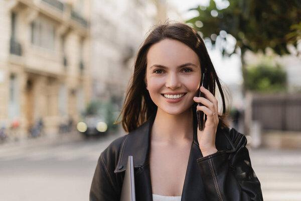portrait of happy woman in black leather jacket holding laptop and talking on smartphone on street in paris
