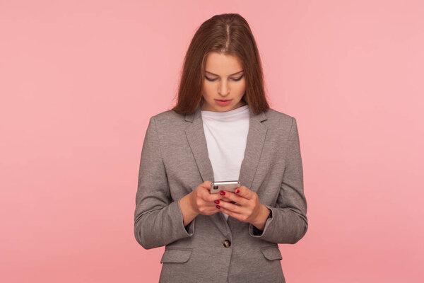 Mobile business application. Portrait of serious focused elegant lady boss in suit reading message, checking news on smartphone, using online services. indoor studio shot isolated on pink background