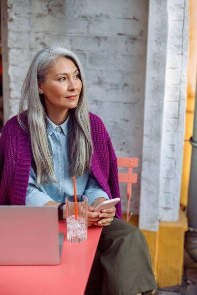 Beautiful mature Asian woman holds mobile phone sitting at coral table with notebook
