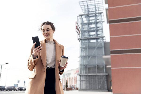 Business Woman smiling charmingly, using her Smartphone. Female enjoying sunny day and funny conversation. Typing on her Phone with Interest. Brunette Girl. Successful Woman. Phones. Apps.