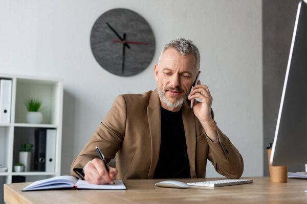 handsome businessman talking on smartphone and writing in notebook in office 