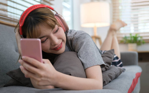 Smiling young woman wearing headphones and using smart phone, resting on couch in cozy living room.