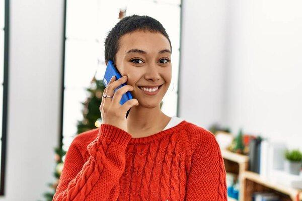 Young hispanic woman talking on the smartphone standing by christmas tree at home