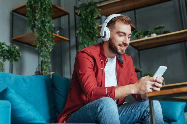 low angle view of happy bearded man listening music in headphones and using smartphone in living room 