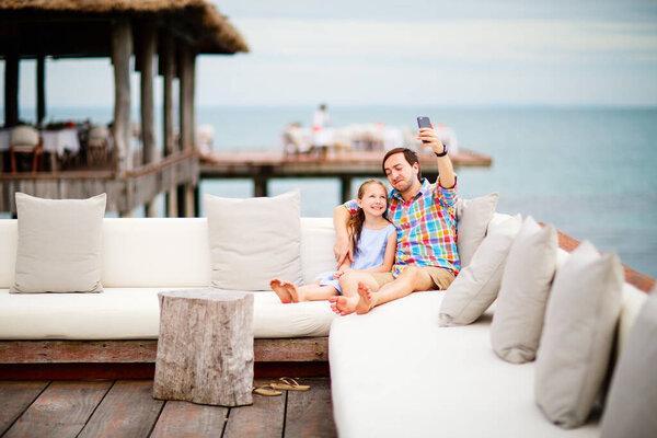 Father and daughter relaxing outdoors at luxury resort taking selfie with smartphone