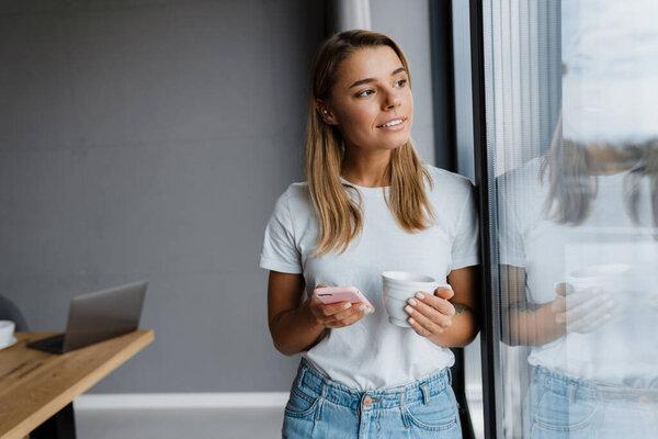 Beautiful blonde girl using mobile phone drinking coffee at home kitchen