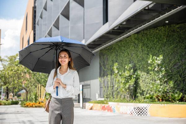 在炎热的阳光下，一位年轻的亚洲女商人拿着雨伞步行去工作。她的严肃态度和职业风度反映了她在商界的决心和成功.
