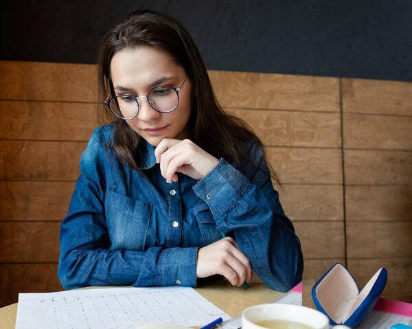 a pensive girl in round glasses sits in a cafe and looking at the drawing, she fills out a lifestyle schedule drawn by her with felt tip pens, portrait, a business photo session