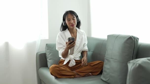 使用手机技术坐在沙发上轻松地微笑着的年轻女性翻阅并欣赏移动应用程序中的在线电子商务购物，观看嵌入式电影和音乐.