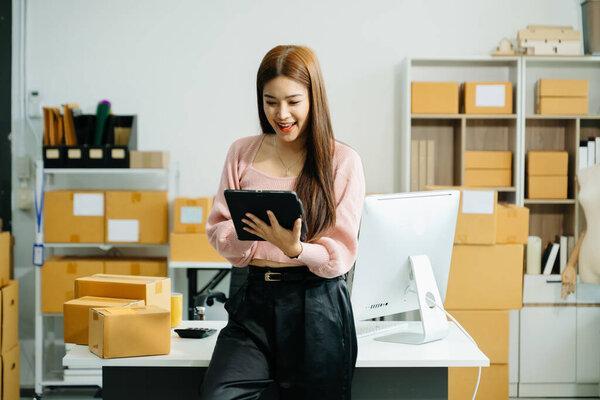 ， 年轻的企业主女人准备包裹盒，， 使用数字平板电脑并检查在线订单以交付给客户。在线购物概念。