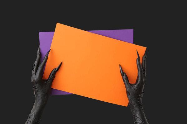 Black hands of witch with blank paper sheets on dark background. Halloween celebration