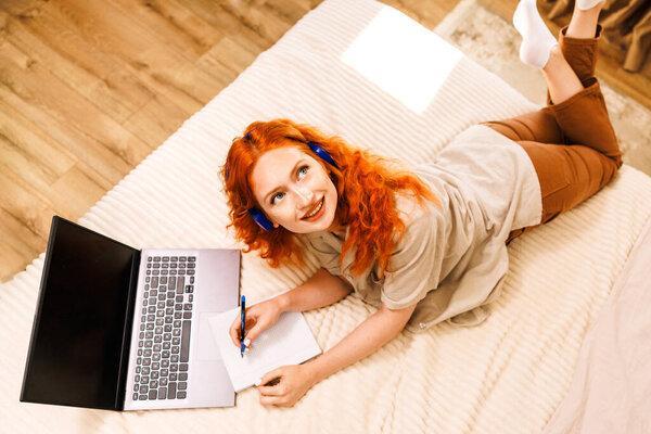 Top picture of a beautiful girl lying on the bed surfing the internet on the computer with headphones on. High quality photo