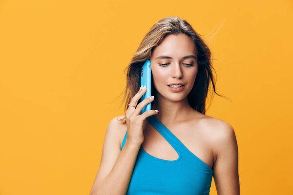 Stylish woman in blue tank top chatting on smartphone in front of vibrant yellow wall