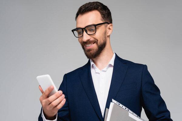 young businessman in blue blazer and eyeglasses looking at mobile phone isolated on grey
