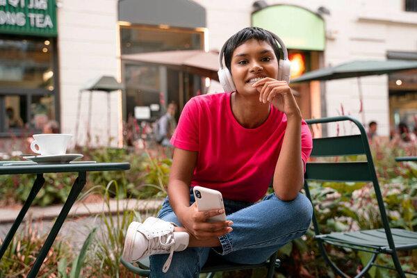 Smiling Indian woman using mobile phone, reading text message, communication online, social media concept. Asian hipster female wearing wireless headphones, listening music on the street
