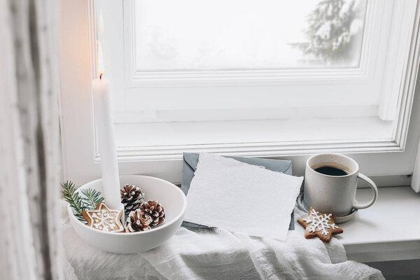 Christmas still life scene. Blank greeting card mockup. Cup of coffee, gingerbread cookies and fir tree branches. Burning candle in the ceramic candle holder. Seasonal photo. Scandinavian interior.