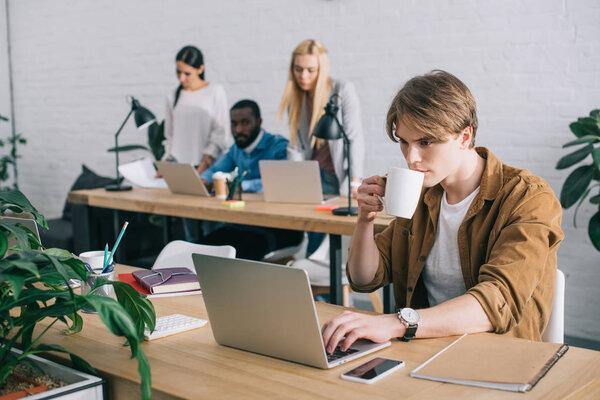 businessman with coffee mug using laptop and colleagues working behind in modern office 