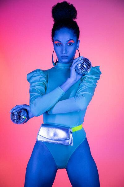 Fashionable african american woman with neon visage holding disco balls on pink background